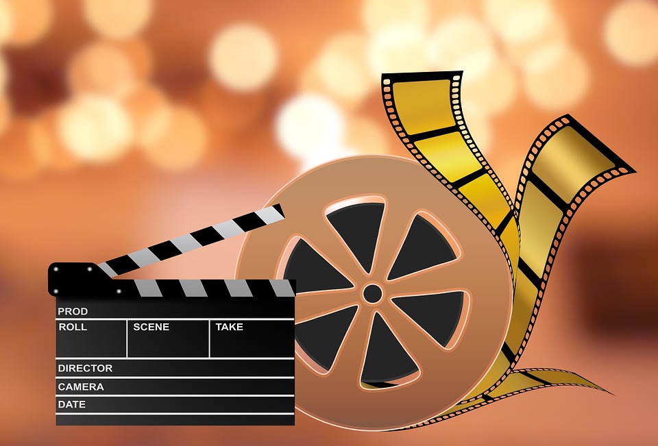 redish orange blurry background with blurry yellow circles to represent lights with a gold bronze movie reel with yellow and black film strips and a black clapperboard 