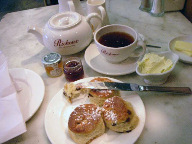a table with a white teapot and a white tea cup and saucer with dark tea inside and a white plate with scones and a knife with butter on it. Also on the table is two small jars of jam and a white bowl with butter. 