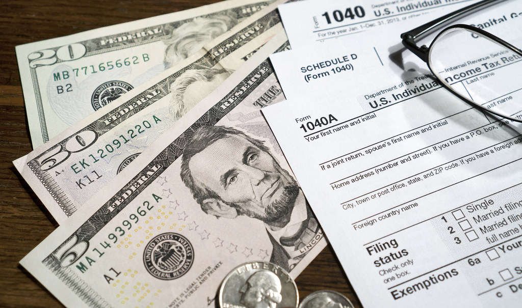 a twenty, a fifty, and a five dollar bill spread like a fan on a table, with a IRS tax form 1040 on top with a pair of glasses on the top of the IRS forms