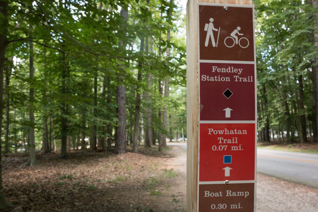 A wooden sign at Pocahontas State Park. The first of three blocks is a brown square with white outlines of stick figure hiking and another biking. The second box is a brown or maroon color and the words Fendly Station Trail with a black diamond and a white arrow pointing forward. The third block is red with the words Powhatan Trail 0.07 mi. a blue square and a white arrow pointing forward. The final box is a brown box with the words Boat Ramp 0.30 mi. In the background of this post/sign are trees 