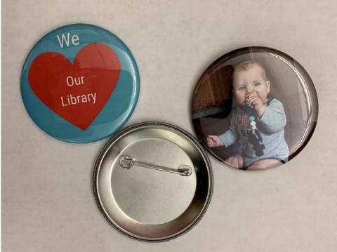 three circles, two of which are buttons, and the third is a magnet. First circle has a blue background with a red heart with white text saying "we love the library". Second circle is the metal pin that is the back of the button that pins to your shirt. The third circle is a picture of a baby