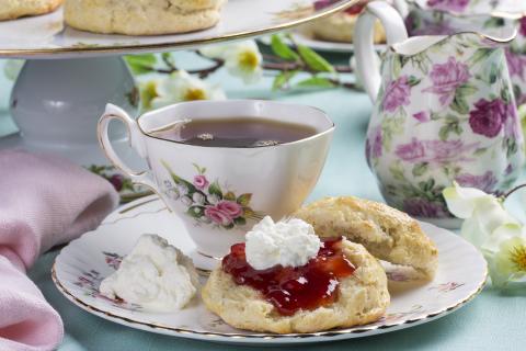 a flowery tea cup and saucer  with gold edges sits on a green tablecloth. The teacup has tea and is sitting on top of the saucer. On the saucer with the cup is a biscuit or scone with jam and cream. There is also a dollop of cream on the side next to the biscuit. There is also a flowery pitcher on the table and scattered around the table are tree blossoms.  