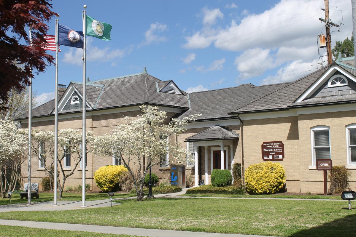 The Dinwiddie Library, a white brick building with blossoming trees out front and three flag poles. The flags displayed are the County of Dinwiddie, the State of Virginia, and the United States Flag